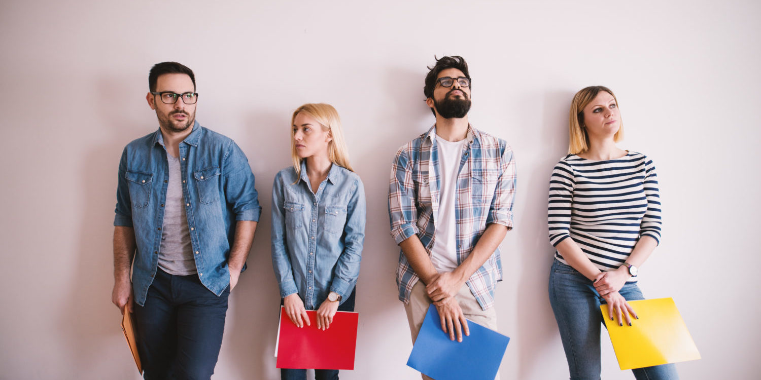 Group of young stylish people leaning against the wall bored before a job interview with folders in hands in the waiting room.